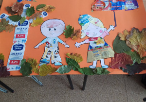 Na stole leży plakat przedstawiający chłopca i dziewczynkę z parasolką oraz drzewo. Wszystkie te elementy sa oklejone etykietami i mlecznych wyrobów.